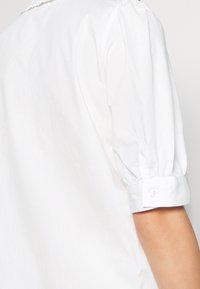 Nærbillede af en hvid skjortes manchette med en enkelt knap, båret på en persons arm mod en lys baggrund.