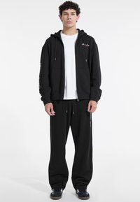 Sort zip-up hoodie med en hvid t-shirt under, parret med sorte sweatpants. Har snoretræk og sidelommer. Enkel design.
