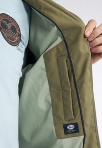 Olivengrøn jakke indvendigt med lysegrøn foering, prydet med et patch og glat tekstur. Inkluderer en sidelomme og brandlabel.