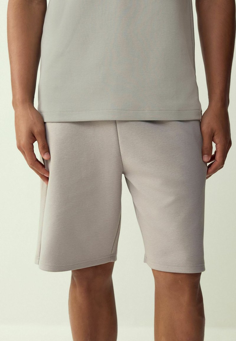 Uomo che indossa pantaloncini beige chiaro semplici a lunghezza al ginocchio e una maglietta abbinata, in piedi con le mani rilassate ai lati contro uno sfondo neutro.