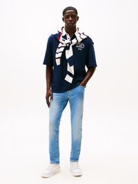 Fiatal férfi világoskék farmernadrágban, sötétkék pólóban a "Tommy Jeans University Club" felirattal, fehér sneakersben és csíkos pulóverrel a vállán.