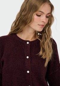 Burgunderfarbener Strickcardigan mit rundem Ausschnitt, ausgestattet mit fünf großen Perlenknöpfen an der Vorderseite und einem weichen, strukturierten Stoff.