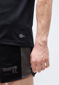 Κοντινό ενός χεριού και μπράτσου δίπλα σε μαύρο σορτς με την επιγραφή «CHAMPION F.C.» και μαύρο αθλητικό μπλουζάκι «C Dry».