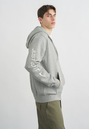 Νέος άνδρας με γκρι φούτερ Nike με κείμενο «Just Do It» στο μανίκι και χακί παντελόνι, όρθιος με τα χέρια στις τσέπες, στραμμένος προς τα δεξιά.