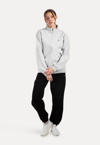 Joven de pie con las piernas cruzadas, vistiendo un sudadera de media cremallera gris claro, pantalones deportivos negros y zapatillas blancas, contra un fondo sencillo.