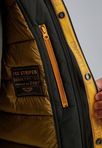 Interno della giacca isolante nera e oro con una cerniera gialla e fodera. Presenta un'etichetta con "ICE STRIPER", numero di emissione e numero di disegno.