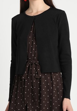 Kobieta w czarnym, krótkim kardiganie na brązowej sukience w groszki, przewiązanej kokardą w talii.