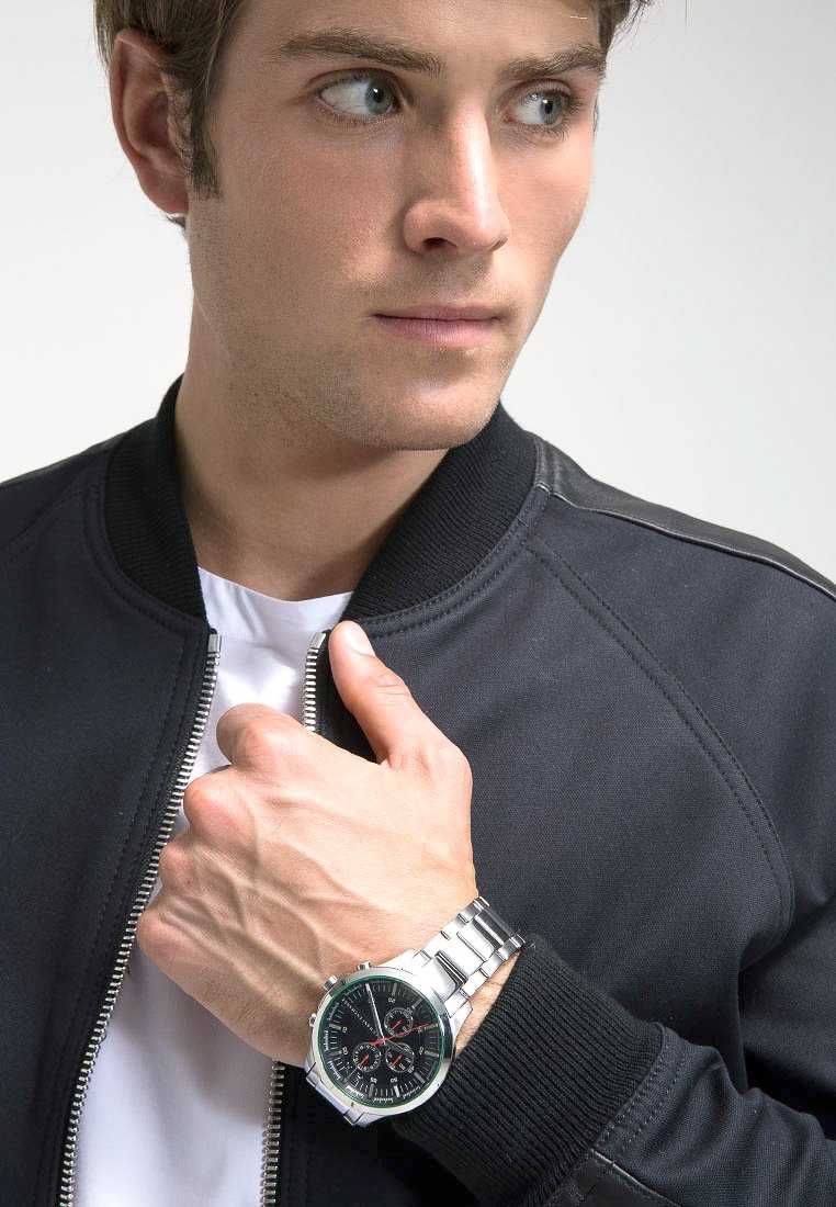 Reloj plateado con una esfera negra y acentos rojos en una correa de eslabones metálicos, llevado en una mano, con una chaqueta bomber negra y una camisa blanca visibles.