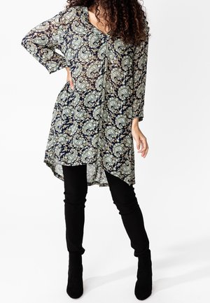 Luźna granatowa tunika z wzorem paisley w zieleni i kremie; ma długie rękawy, dekolt w serek oraz asymetryczny dół, zestawiona z czarnymi spodniami.