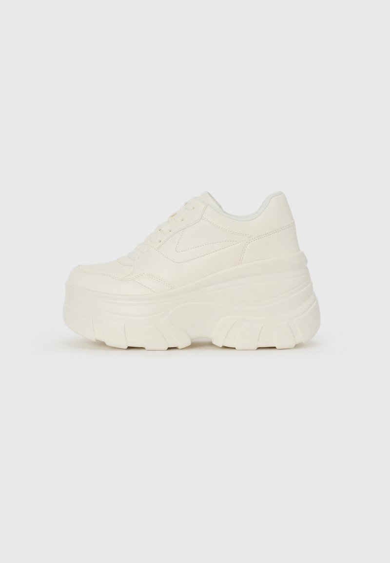 Sneaker à semelle épaisse style plateforme en blanc cassé uni avec lacets à l’avant et semelle épaisse texturée sur un fond clair uni.