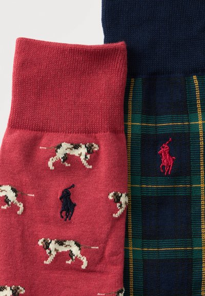 Gros plan de deux chaussettes pliées, une rouge avec des motifs de chiens et un logo joueur de polo bleu marine, l'autre à carreaux bleu marine et verts avec un logo joueur de polo rouge.