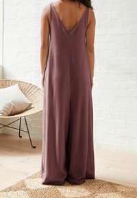 Une combinaison de couleur prune avec un dos en V, des jambes larges et un tissu doux. Elle se caractérise par des détails minimaux et des poches latérales.