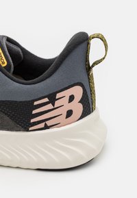 Grå sneakers med en teksturert overdel, rosa "NB"-logo, rund hvit såle og en hælflik med gul aksent.