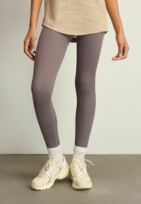 Lette lilla ribbede leggings med en tætsiddende design. Båret med beige sneakers og hvide sokker mod en neutral baggrund.