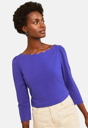 Woman with short curly hair wearing a purple top with scalloped neckline and sleeve edges, paired with light beige pants.