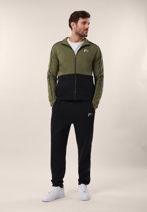 Homme portant une veste zippée verte et noire et un jogging noir avec des baskets blanches, debout les mains dans les poches, regardant vers la droite.