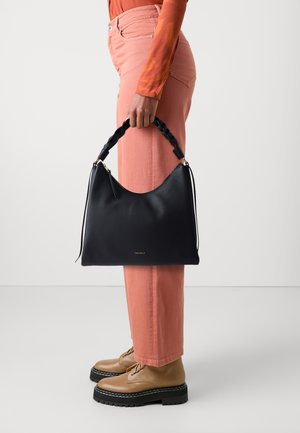 Borsa a tracolla in pelle nera con manico intrecciato, caratterizzata da una texture liscia e un sottile marchio dorato, tenuta accanto a pantaloni rosa chiaro e scarpe marroni.