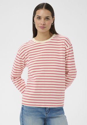 Žena s dlhými tmavými vlasmi, ktorá nosí zlaté kruhové náušnice, krémovú blúzku s dlhými rukávmi s ružovými horizontálnymi pruhmi a svetlomodré džínsy.