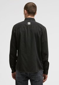 Mann von hinten gesehen, der ein schwarzes, knopfgebundenes Hemd und dunkle Jeans trägt, vor einem schlichten hellgrauen Hintergrund.