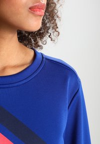 Blå sportströja med ribbat runt halsen, med horisontella ränder i röd och mörkblå över bröstet. Slät tygtextur.