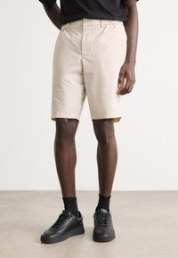 Beiga shorts med slät front, bälteshål och fickor, som bärs med svarta sneakers och strumpor. Materialet verkar slätt och lätt.