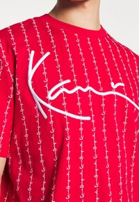 Röd bomullst-shirt med vita broderade ankare och "Kani"-logotyp i kursiv, med rundad halsringning och korta ärmar.