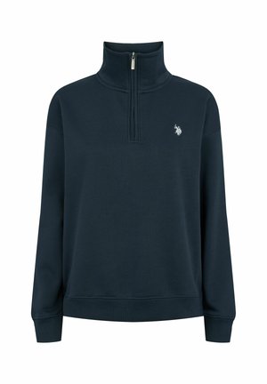Kvinde iført en marineblå half-zip sweatshirt med hvidt broderet logo på brystet og mørkeblå jeans, står med afslappede arme.
