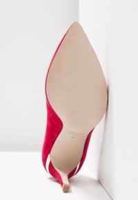 Chaussure à talon haut en daim rouge à bout pointu, inclinée contre un mur blanc, montrant la semelle beige avec la taille 37 et l'inscription Made in Italy.