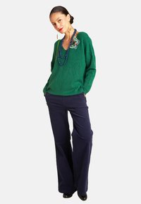Grüner Strickpullover mit V-Ausschnitt und dekorativem Patch, kombiniert mit weit geschnittenen marineblauen Hosen. Der Stoff hat einen leichten Glanz und eine weiche Textur.