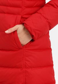 Röd dunjacka med vadderat mönster. Har en sidoficka och elastiska muddar. Materialet verkar slätt och isolerande.