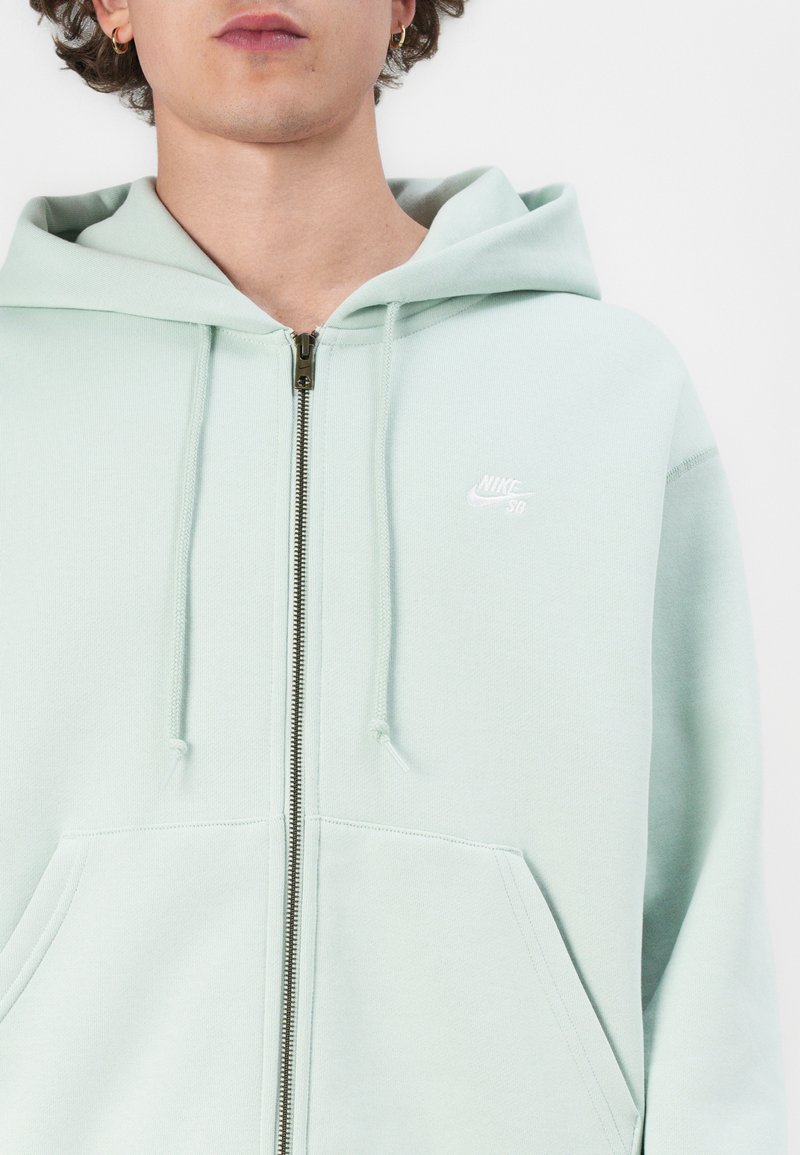 Jasnozielona bluza Nike na zamek wykonana z miękkiego materiału, z przednią kieszenią, kapturem ze sznurkiem oraz małym białym logo na lewej piersi.
