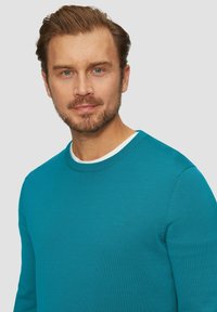 Tyrkysový pletený sveter s okrúhlym výstrihom, rebrovanými manžetami a jemným logom na hrudi. Hladká textura, priliehavý strih.
