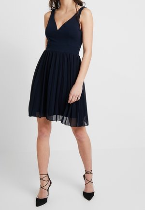 Marineblaues Kleid mit tailliertem Oberteil und plissiertem Rock. Verfügt über einen V-Ausschnitt und dünne Schulterträger. Wird mit schwarzen spitzen Schuhen getragen.