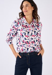 Camicia a maniche lunghe con bottoni caratterizzata da un motivo di foglie colorato nei toni del rosa, blu navy e bianco, realizzata in tessuto leggero. Abbinata a pantaloni in denim scuro.