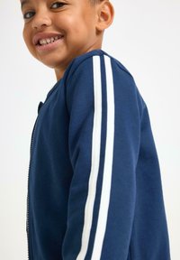 Marine blauwe sweatshirt met rits, gekenmerkt door witte dubbele strepen op de mouwen, gemaakt van een zachte stof met een gladde textuur en een casual pasvorm.
