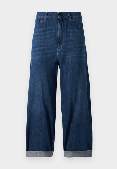 Calças de ganga largas de cor azul-escura com bainhas dobradas, botão frontal e bolsos, contra um fundo claro e liso.