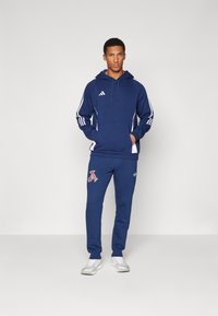 Mornarsko moder pulover s kapuco z belimi črtami in kenguru žepom; ujemajoči se trenirke z logotipom; v kombinaciji z belimi atletskimi čevlji.