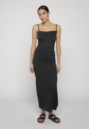 Femeie în picioare, purtând o rochie midi neagră fără mâneci, cu bretele subțiri și sandale negre cu cataramă, pe un fundal simplu și deschis la culoare.