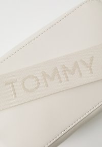 Barna színű csuklós táska, texturált szövetpánttal, amelyen bemélyített "TOMMY" felirat található, sima szintetikus anyag és kontrasztos varrási részletek.