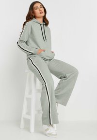 Conjunto de sudadera gris claro y pantalones de pierna ancha con franjas laterales negras. La sudadera tiene un cordón y un bolsillo tipo canguro; está hecha de una tela suave.