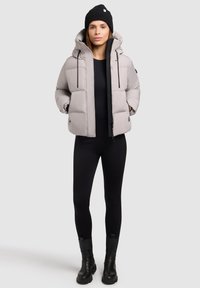 Lys grå pufferjakke med hætte, der har lynlås foran, elastiske snore og sidelommer. Båret over et sort tætsiddende outfit og sorte støvler.