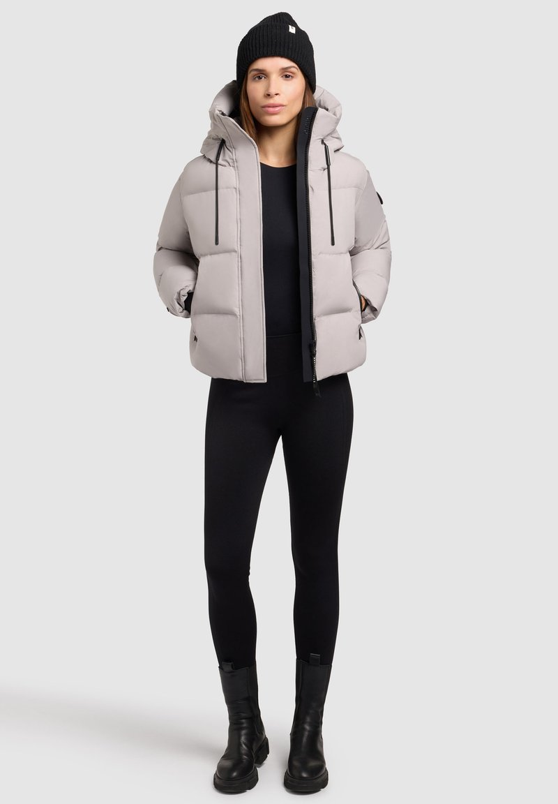 Lys grå pufferjakke med hætte, der har lynlås foran, elastiske snore og sidelommer. Båret over et sort tætsiddende outfit og sorte støvler.