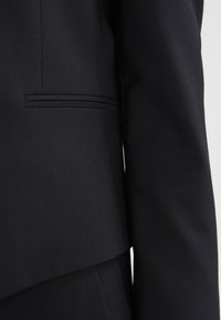 Svart skräddarsydd kavaj i texturerat tyg, med strukturerad slag och en diskret jettficka, med en slank och modern design.