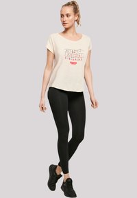 Light beige short-sleeve t-shirt with graphic text design, paired with black leggings and black athletic shoes. Smooth, casual fabric.