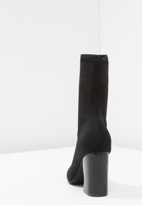 Botte chaussette noire montante jusqu'au genou avec un talon haut épais et brillant, vue de dos sur une surface blanche avec un fond clair uni.