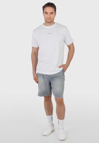 Homme portant un t-shirt blanc à manches courtes avec un petit logo, un short en denim gris, des chaussettes blanches montantes et des baskets blanches, debout avec une main dans la poche.