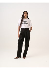 Bílé tričko s černým grafickým textem, zkombinované s černými kalhotami s vysokým pasem. Model má na sobě špičaté hnědé boty; minimalistický design.