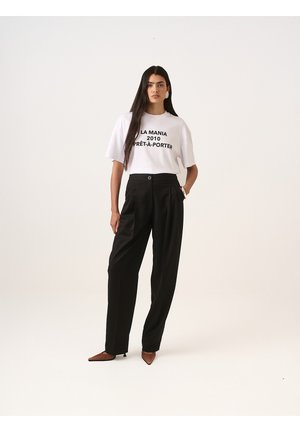 Bílé tričko s černým grafickým textem, zkombinované s černými kalhotami s vysokým pasem. Model má na sobě špičaté hnědé boty; minimalistický design.