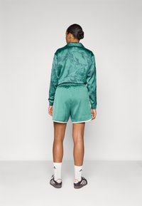 Casaco de treino verde em satin com padrão de folhas, combinado com calções verdes sólidos com uma bainha branca. O modelo usa meias brancas até ao tornozelo e sapatilhas pretas.