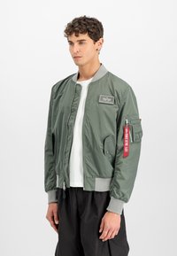 Olive green bomber jacket with a silver-tone zipper, ribbed cuffs, and waistband. Features a patch on the chest and a pocket on the sleeve.